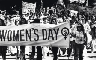 Women marching holding a white banner that says "Women's Day" and has the female symbol after it