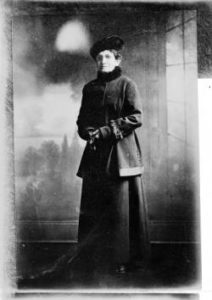 Standing portrait of women's suffragist Minnie Reynolds Scalabrino in Denver, Colorado; she wears a wool coat with a fur collar and cuffs, gloves, and a hat.