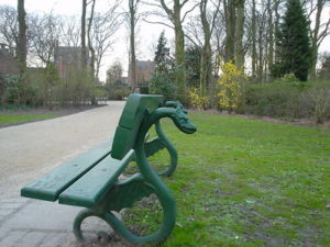 A green bench with two wooden planks sits along a park path in Belgium. The legs/back of the bench are shaped like dragons.