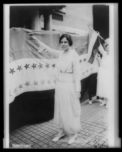 Alice Paul, full-length portrait, standing, facing left, raising glass with right hand