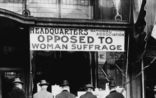 Sign over a storefront that reads "Headquarters National Association Opposed to Woman Suffrage" with four men appearing to read papers while facing the storefront. A woman looks back at them as she walks by. Circa 1911.