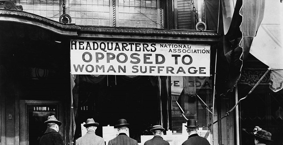 Sign over a storefront that reads "Headquarters National Association Opposed to Woman Suffrage" with four men appearing to read papers while facing the storefront. A woman looks back at them as she walks by. Circa 1911.
