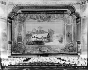 Elitch Garden's original summer stock theater curtain, West Thirty-eighth Avenue, Denver, Colorado, with proscenium and decorative scene painted on curtain; shows English side gable cottage with walled fence garden; quote at bottom of curtain: "Ann Hathaway's Cottage, a mile away, Shakespeare sought at close of day." Rows of wooden theater seating and a piano in foreground.