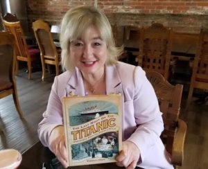 Author Veronica HInke sits at a wooden table holding up a copy of her book"Last Night on Titanic"