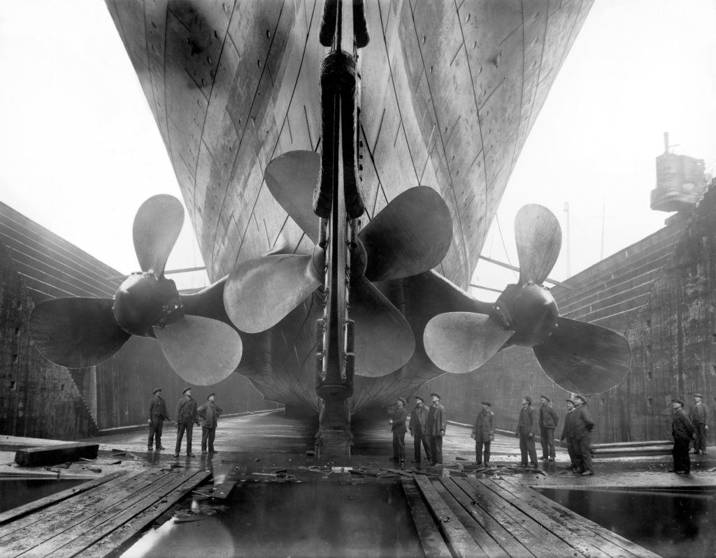 A group of men stand below three giant propellers underneath the bow of the Olympic in dry dock.