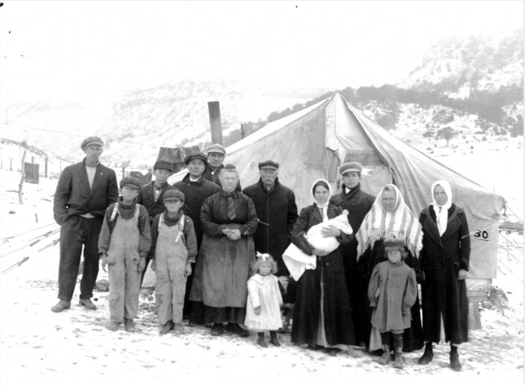 A family of miners, mostly women and children, stand in front of a tent. Snow covers the ground.