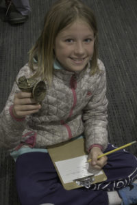 A young girl smiles up at the camera while holding an object up to see. A clipboard is in her lap and a pencil is in her hand, apparently taking notes.