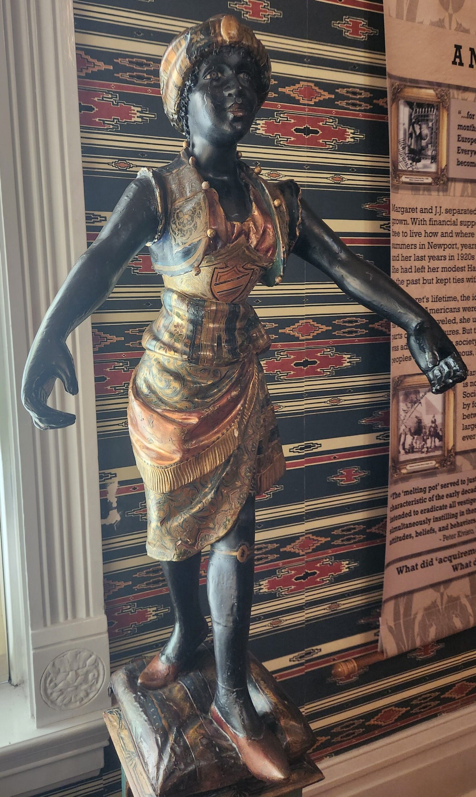 20250409_105149 A blackamoor statue of a young woman. Her arms are extended in front of her and she is wearing a red, blue, and gold turban and dress.