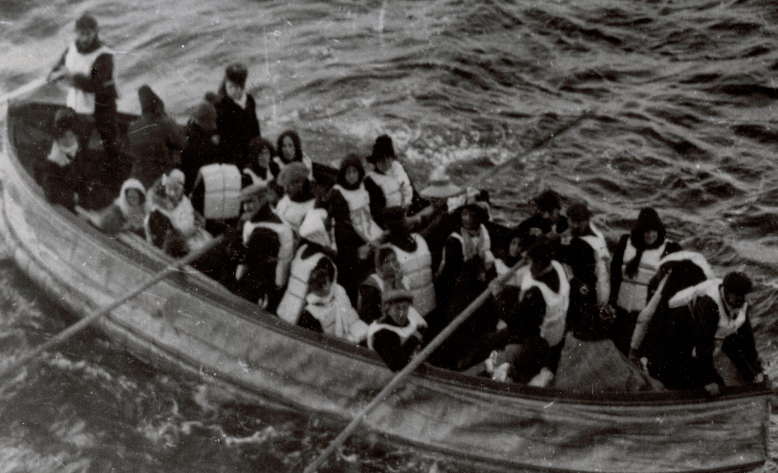 Titanic Lifeboat transfer from #14 National Archives A black and white photograph of a lifeboat at sea. It is full of people wearing lifeboats, with some rowing.