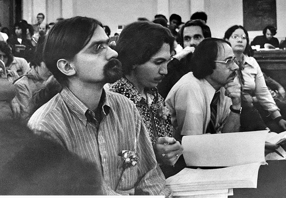 Black and white photo of people attentively watching something, seated and holding documents.