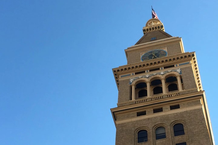 Tall brown brick tower with clock face against clear blue sky.
