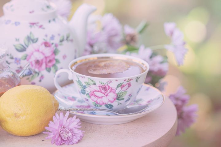 Floral teacup and teapot with tea, lemon, and flowers on a table.