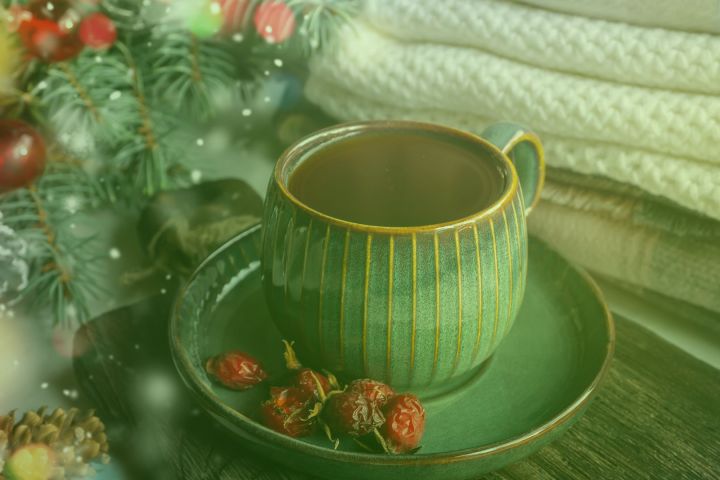 Green cup and saucer with dried berries near folded blankets and pine branches.