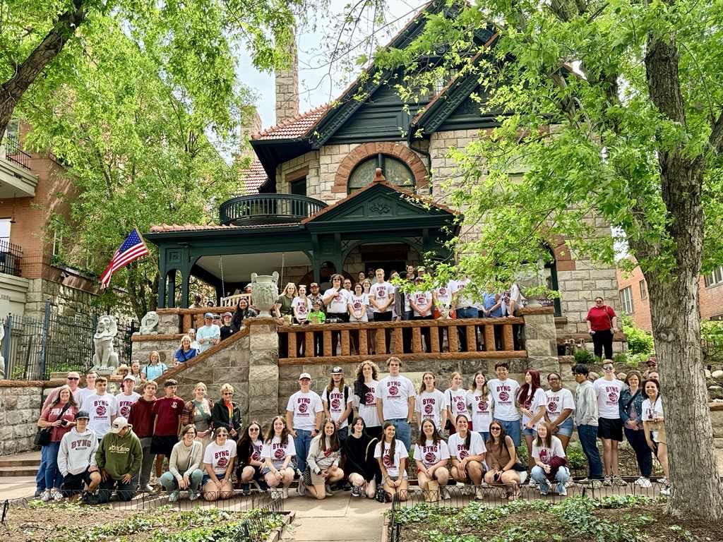 Large group in matching shirts at historic stone house with porch, surrounded by trees.
