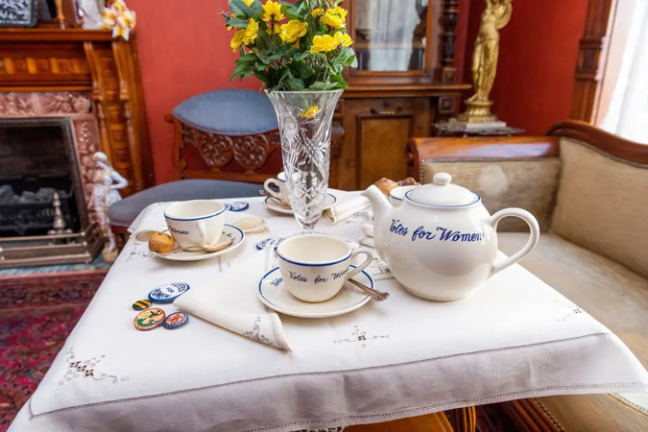 Vintage tea set with 'Votes for Women' on table, vase with yellow flowers, badges, and a pen in a cozy room.